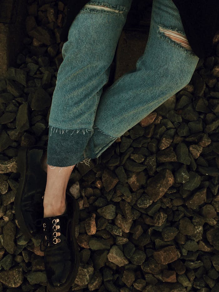 services-01 Close-up of distressed denim jeans and black shoes on a rocky ground, showcasing urban fashion.